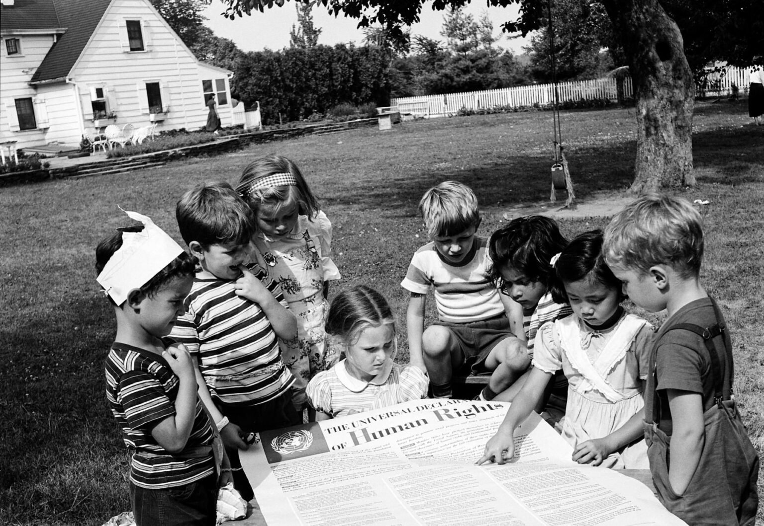 Un grupo de niños pequeños, de pie en el césped, leyendo un póster grande de la Declaración Universal de los Derechos Humanos.