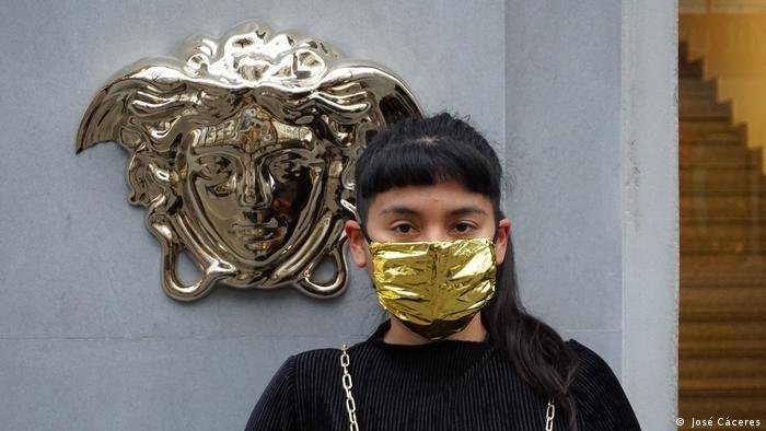 Artista con mascarilla dorada frente al logo de Versace en el Museo Ludwig.