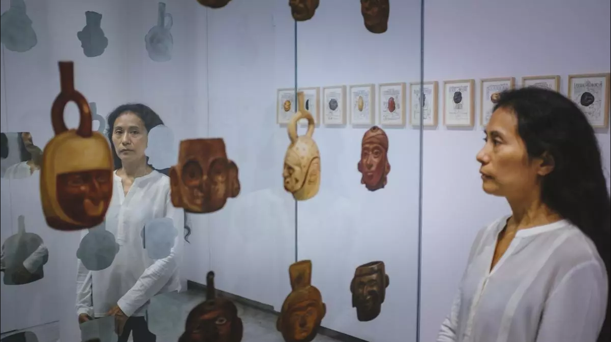 Una mujer observa una instalación artística con réplicas de objetos precolombinos y documentos enmarcados, en una exposición sobre la hispanidad.