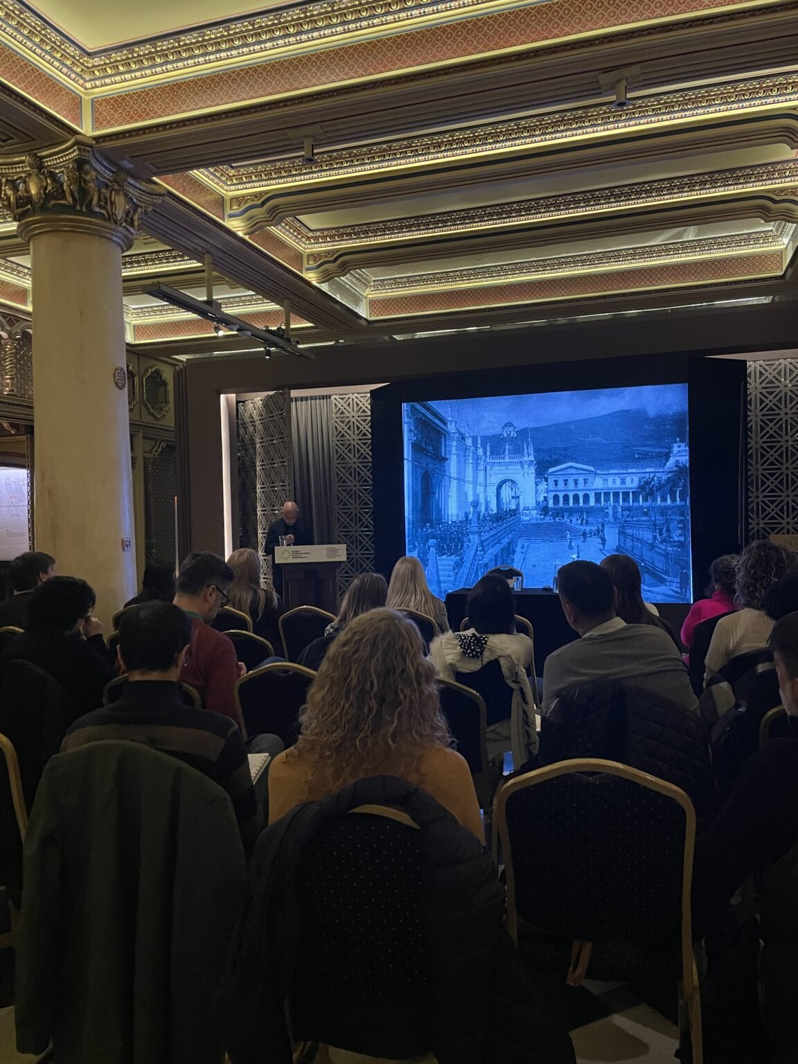 Público asistiendo a una presentación sobre museos, con una imagen antigua proyectada en una pantalla.