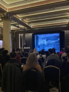 Público asistiendo a una presentación sobre museos, con una imagen antigua proyectada en una pantalla.